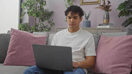 Young hispanic man using laptop on sofa in cozy living room interior