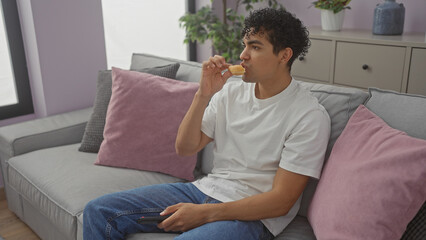 A young hispanic man relaxes on a sofa in a modern living room, casually dressed, snacking and holding a smartphone.