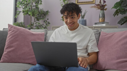 A cheerful young hispanic man using a laptop on a cozy couch in a modern living room setting