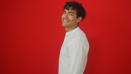 A smiling handsome young hispanic man in a white shirt against an isolated red background wall