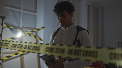 A young hispanic man analyzes evidence on a tablet at an indoor crime scene with 'do not cross' tape