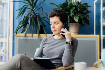 Confident Neutral gender middle aged woman in casual clothing having conversation by phone, making note in notebook in office, meeting room. Phone interview. HR specialist, hiring, recruiting
