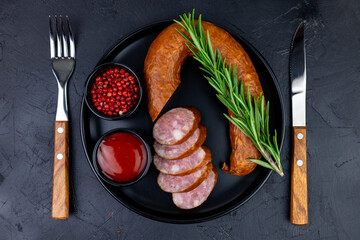 Sausage salami pieces on a black plate with spices with a fork and knife top view