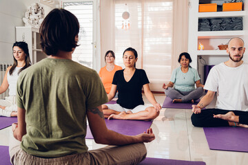 Group of people in a yoga class. Women and men doing yoga on the purple mat in the lotus position with the trainer in front.