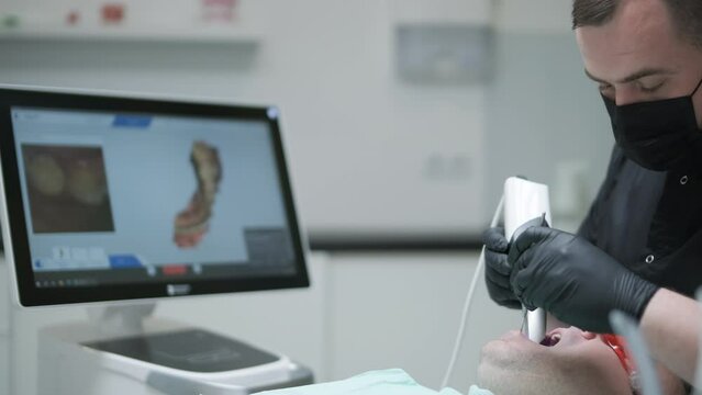 Doctor scans the patient's teeth in the clinic. The dentist holds in his hand a manual 3D scanner for the jaw and mouth. Dental health. Creates a 3D model of teeth and gums on a medical monitor.