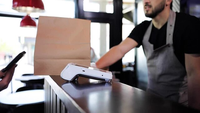 Close up of a feminine hand holding a smartphone with an NFC payment technology used for paying for take away meal in a cafe. Customer uses mobile to pay for food through a credit card terminal
