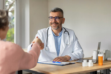 Older female patient feeling gratitude to happy male GP doctor, shaking hand of therapist at appointment in office, thanking for medic care, therapy, support