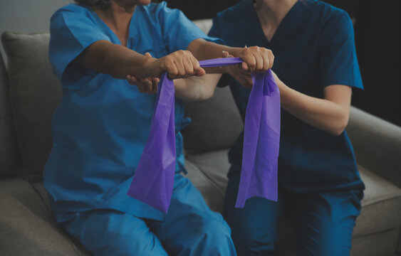 Personal trainer assisting senior woman with resistance band. Rehabilitation physiotherapy worker helping old patient at nursing home. Old woman with stretch band being coached by physiotherapist.