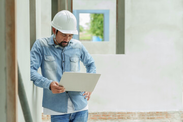 Asian male engineer with hard hat using laptop at construction site. Concrete wall background....