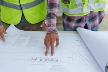 Close-up of two Asian male engineers with safety vests pointing at blueprints at construction site....