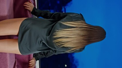A woman with long blond hair in a black satin robe stands by an open window and looks at the evening sky. Vertical view.