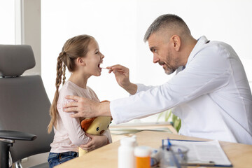 Obraz premium Attentive doctor. Determined professional male doctor examining little girl patient and looking in her throat during checkup