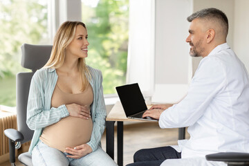Male gynecologist doctor using laptop computer and talking to pregnant European woman at hospital....