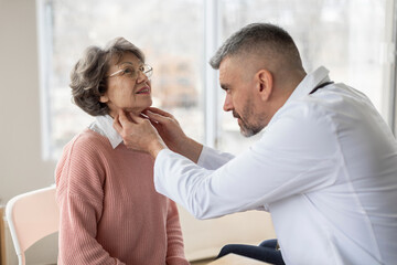 Professional male doctor palpating neck of senior female patient while examining her during consultation in private clinic office