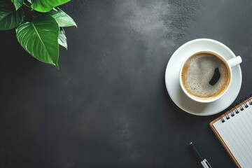 Overhead view of a fresh cup of coffee beside a notebook and a green plant on a dark surface