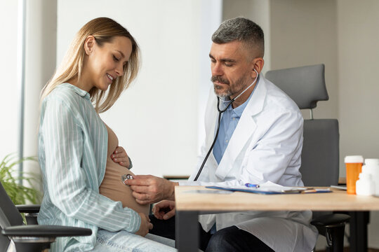 Pregnant woman having appointment with male doctor at clinic, gynecologist OB GYN medic specialist with stethoscope listens to baby's heartbeat in mother's belly