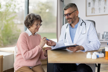 Happy male doctor consulting senior woman, friendly professional physician talking to elderly woman signing medical paper during appointment visit in clinic.