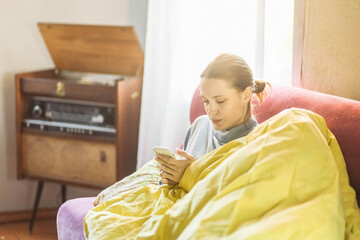 beautiful woman on the sofa under a blanket using a smartphone on a sunny day