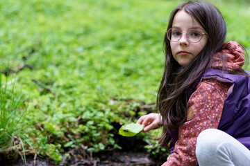 A small beautiful smiling long-haired dark-haired girl in glasses and a vest sits near a stream in the forest