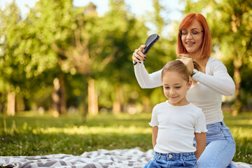 Photo of a ginger-haired mom using a comb and styling her daughter's hair at the park, sitting on a...