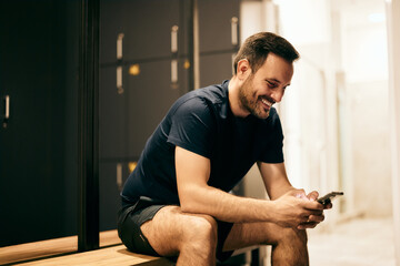 The smiling man sitting on a bench in the locker room at the gym and using a mobile phone.