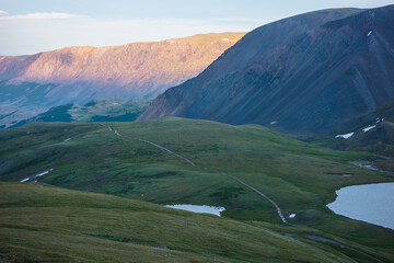 Twilight aerial view to long dirt road between two alpine lakes on hills against sunlit mountain range of gold sunset color. Scenic landscape with mountain lake in shadow and golden rocks at sunset.