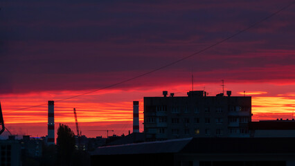 Naklejka premium Urban Sunset. A dramatic sunset sky over an urban landscape with silhouettes of buildings and industrial elements against a vibrant backdrop of red and orange hues.