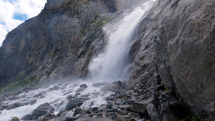 A majestic waterfall cascades down a rugged, rocky cliffside, surrounded by steep, craggy mountains. The powerful flow of water creates a misty spray, adding to the dramatic natural landscape.