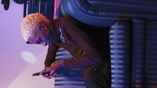 Trandy stylish african american man laying and resting on vintage soft sofa, texting on smartphone. Adult attractive cheerful young creative guy looking at phone screen, playing video games online.