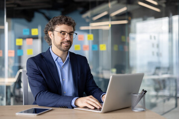 Happy smiling businessman at work with laptop, man in business suit typing on keyboard smiling happily, financier preparing report.