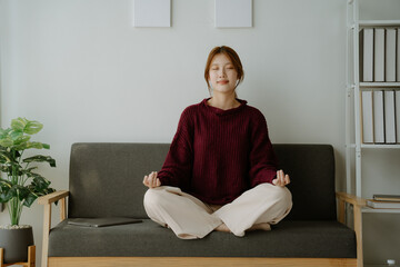 Clam of Asian young woman doing yoga lotus pose to meditation and relax on couch during work online...