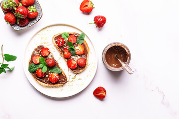 Sweet homemade toasts with chocolate paste, nuts, srtawberry and aromatic mint leaves on the serving plate on white marble background. Tasty summer dessert.