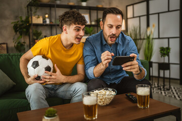 Two excited men friends brother use phone to watch football match home