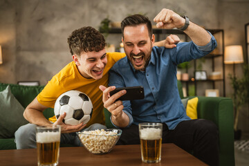 Two excited men friends brother use phone to watch football match home