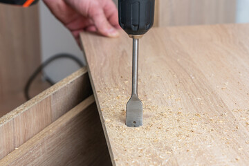 A worker using a drill with a feather drill makes a hole in a wooden slab