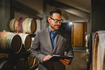 Adult winemaker hold clipboard check barrels with wine in the cellar