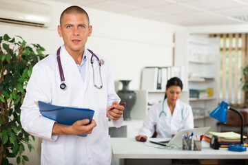 Portrait of polite doctor wearing white coat meeting patient in clinic office, filling out medical...