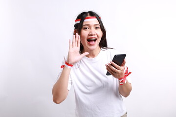 Naklejka premium Young beautiful woman wearing red white flag ribbon holding her phone while shouting and screaming loud with a hand on her mouth. Indonesia's independence day concept.