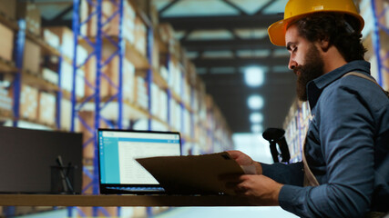 Delivery service associate examines order details on files, working in industrial storage room to...