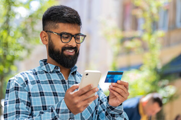 Indian bearded man using credit bank card smartphone while transferring money, purchases online...