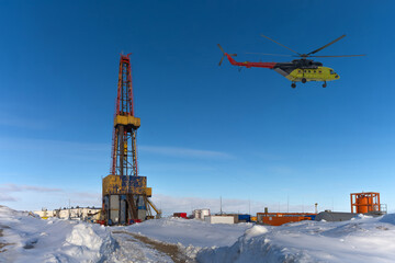 Drilling derrick and well drilling infrastructure in the northern oil and gas field. Polar snowy winter. Yellow helicopter carrying drillers. Clear blue sky. Collage © mangz