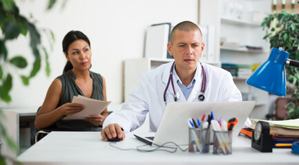 Focused doctor sitting at table with laptop, consulting female patient and writing prescription at clinic..