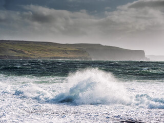 Fototapeta premium A rocky shoreline with a large wave crashing against the rocks. The sky is cloudy and the water is choppy. Cliff in the background. Doolin area, Ireland. Popular tourist area.