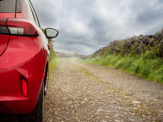 A red car is parked on a small country road with a view of stone fence and the sky. The car is wet...