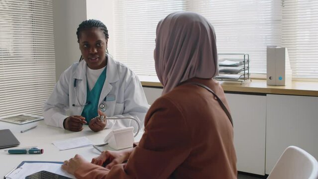 Medium shot of Black female physician sitting at desk in clinic, measuring blood pressure for Muslim patient in Islamic headscarf, while asking questions about symptoms, complaints and medical history
