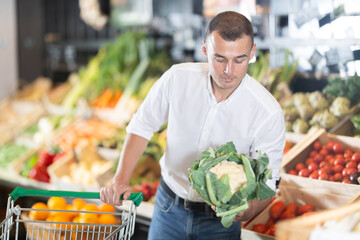Careful young male customer buying fresh cauliflower in grocery store with large assortment