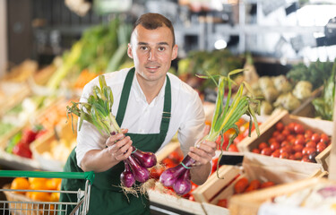 Sales man works in vegetable store bio supermarket and goes through goods. He holds onion in hands, examines it, checks quality
