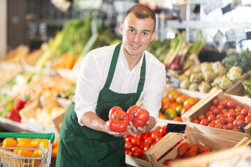 Friendly male seller in green apron enthusiastically offering fresh ripe tomatoes in organic...