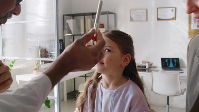 Medium close-up of hands of mature Asian male physician examining girls eyesight, testing movement and reflexes, asking to follow moving pen, giving thumb up and high five, at checkup in GP clinic