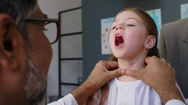Close-up rear shot of mature Asian male physician examining and palpating throat, neck of 7-year-old Caucasian girl, asking to open mouth, then giving high five, during checkup after flu at GP surgery